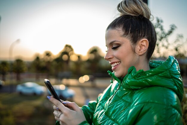 Image of excited young blond Latin woman happy using cellphone on park background