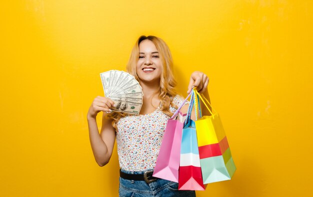 Image of a excited young blonde girl holding banknotes and shopping bag over yellow background