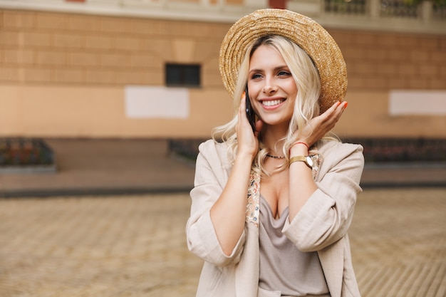 Image of excited young cute woman walking outdoors talking by mobile phone.