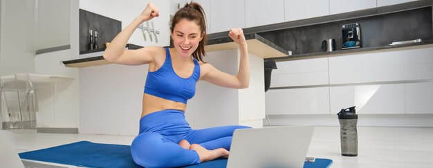 Image of excited young woman showing muscles flexing biceps and looking at laptop screen workout