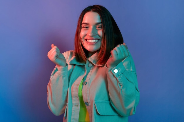 Image of extremely happy Caucasian woman wearing stylish jacket standing with clenched fists celebrating her victory and success posing isolated on neon light background