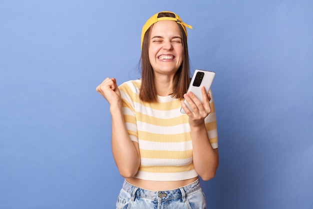 Image of extremely happy smiling teen girl wearing baseball cap and casual Tshirt using cell phone reading message with excellent news clenched fists standing isolated over blue background