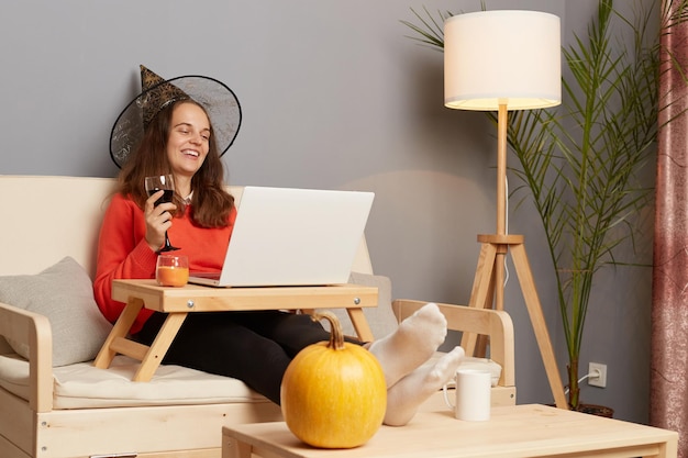 Image of extremely happy woman wearing jumper and witch hat sitting on sofa and using laptop for online conversation drinking wine from glass and laughing happily while celebrating halloween