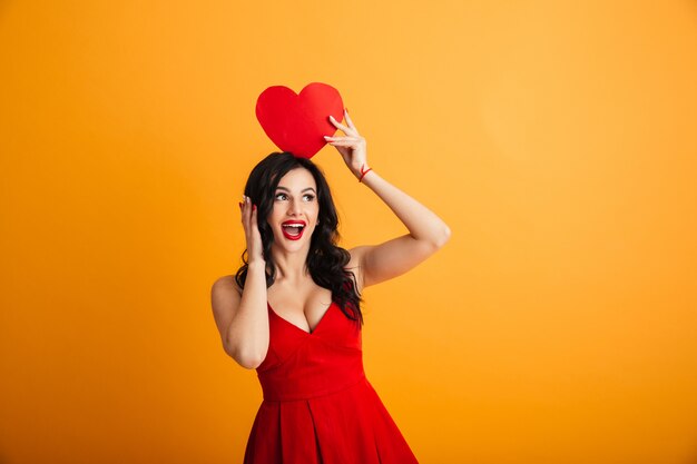 Image of fascinating woman 20s in red dress smiling and holding paper valentine heart above head, isolated over yellow wall