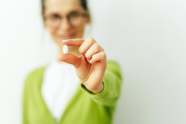 Foto immagine della pillola focalizzata in mano con vista sfocata della donna sorridente bella giovane donna che indossa un cardigan verde e una maglietta bianca che prende la pillola isolata sul muro bianco integratori alimentari vitaminici