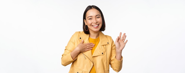 팔을 들고 있는 세련된 노란색 코트를 입은 친근한 아시아 소녀의 이미지는 흰색 배경 위에 서서 인사하며 손을 흔들며 인사하는 자신을 소개합니다