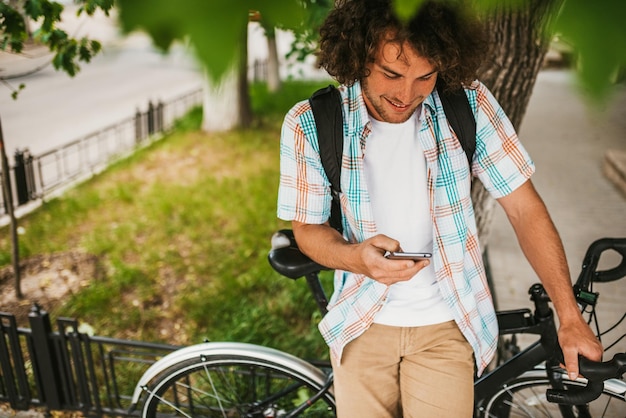 image from above of happy young male student smiling with curly hair wearing shirt with backpack sitting on the bike on the street relaxing texting messages on smart phone people and education
