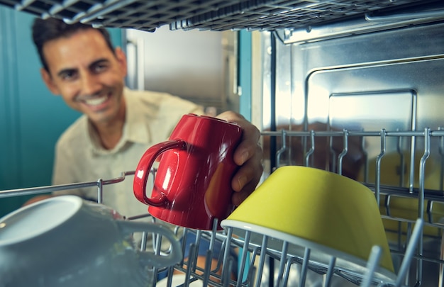 Image from inside a dishwasher of a young man smiling out of focus picking up a red mug next to a yellow bowl in the foreground