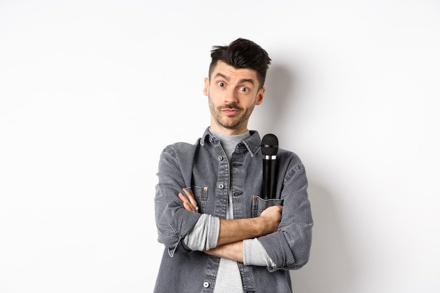 Изображение смешного артиста или певца мужского пола, держащего микрофон в кармане пиджака и смотрящего на камеру со скрещенными руками, стоящего на белом фоне.