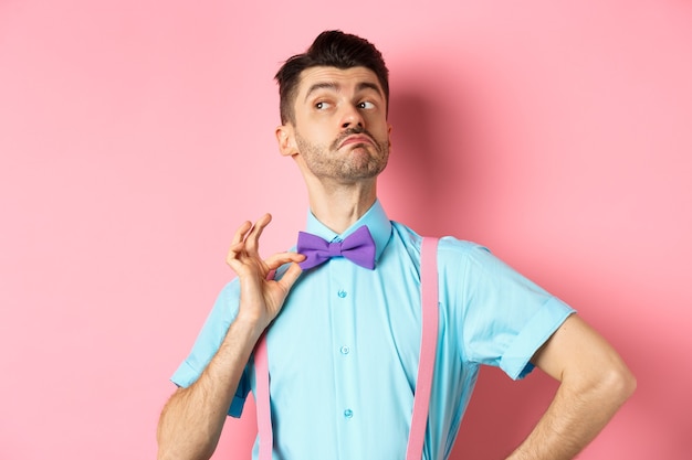 Image of funny man raising chin up and adjusting bow-tie, standing confident and sassy on pink, looking left.