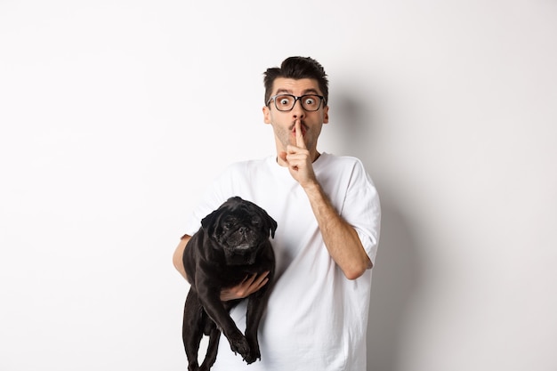 Image of funny young man holding cute black pug dog and shushing at camera, showing hush sign and staring at camera, keeping a secret, standing over white.