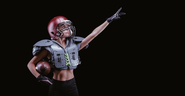 Image of a girl in the uniform of an American football team player. She points to an empty place. Black background. Sports concept. Shoulders pads. Mixed media