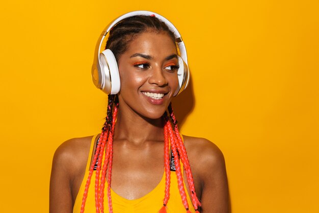 노란색 벽에 격리된 헤드폰으로 웃고 음악을 들으며 아프리카 머리띠를 한 멋진 아프리카계 미국인 여성의 이미지