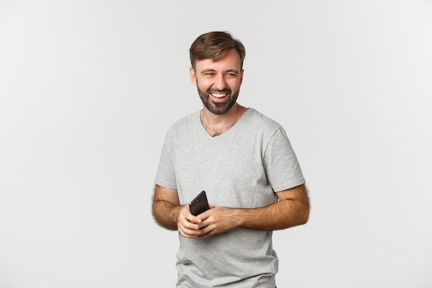 Image of handsome bearded man in gray t-shirt, holding smartphone, looking right and laughing