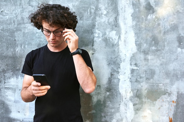 Image of handsome businessman standing outdoors texting on smart phone Young male with curly hair wears spectacles resting outside in the city browsing on his cell phone on concrete gray background