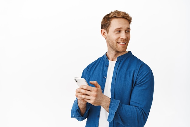 Image of handsome caucasian man using smartphone holding mobile phone in hands and turn head right looking at logo banner with company advertisement standing over white background