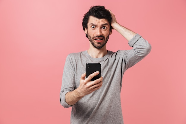 Image of a handsome confused serious young man over pink wall using mobile phone.