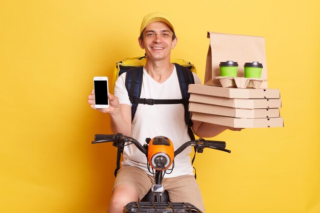 Изображение красивого курьера, показывающего пиццу в картонной коробке, кофе с собой и смартфон с пустой областью для рекламы, сидящего на быстром мотоцикле, изолированном на желтом фоне