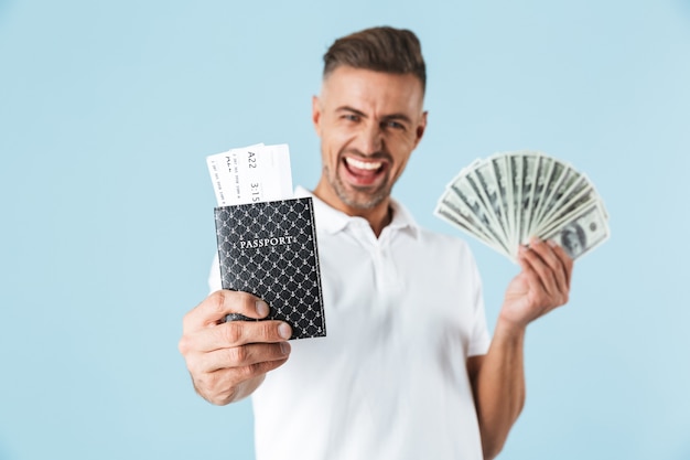 Image of a handsome excited emotional adult man posing over blue wall holding passport with tickets and money.