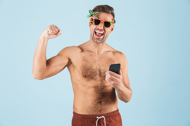 携帯電話を使用して青い壁の上でポーズをとる水着のハンサムな興奮した幸せな大人の男性の画像は、勝者のジェスチャーを作ります。
