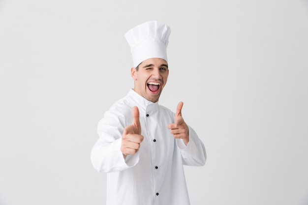 Image of handsome excited young man chef indoors isolated over white wall background pointing to you.