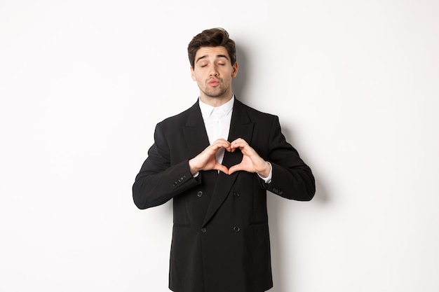 Image of handsome groom in black suit, showing heart sign, close eyes and pucker lips, waiting for kiss, standing against white background.