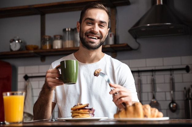 Image of handsome guy 30s sitting at table and eating food while having breakfast in modern apartment