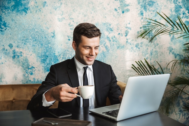 Image of a handsome happy young businessman sitting in cafe using laptop computer drinking coffee.