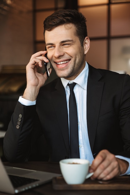 Image of a handsome happy young businessman sitting in cafe using laptop computer talking by mobile phone.