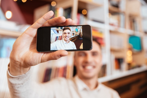 Foto immagine di un bel giovane felice vestito in camicia seduto al bar e fare selfie con il suo telefono.