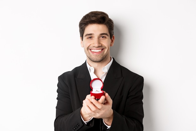 Image of handsome man looking romantic, open small box with engagement ring, making a proposal and smiling, standing against white background.