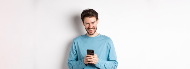 Изображение красивого мужчины, использующего смартфон и смеющегося, улыбающегося в камеру, радостно стоящего над белым пляжем