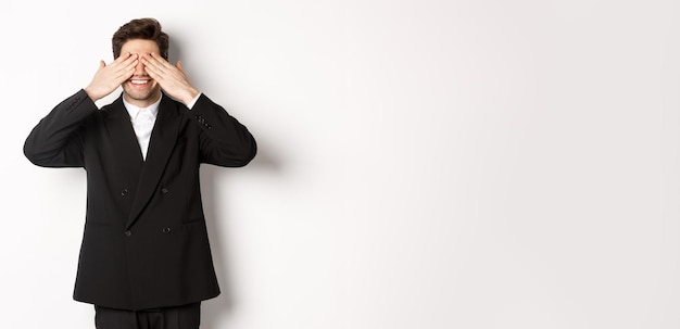 Photo image of handsome stylish man in black suit waiting for christmas surprise covering eyes with hands