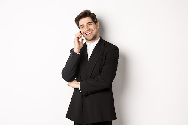 ハンサムで成功したビジネスマンが電話で話し、満足して笑って、白い背景にスーツを着て立っている画像