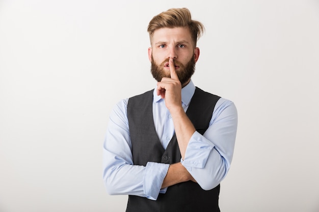 Image of a handsome young bearded man standing isolated over white wall make silence gesture.