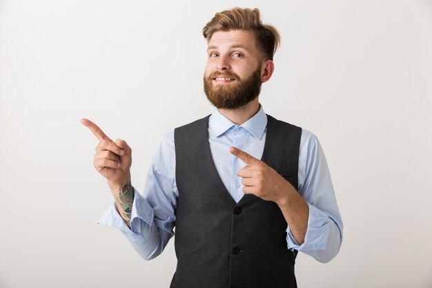 Image of a handsome young bearded man standing isolated over white wall pointing.