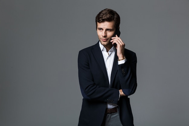 Image of a handsome young business man isolated over grey wall wall talking by mobile phone.