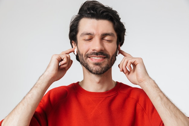 Image of a handsome young excited man posing over white wall listening music with earphones.