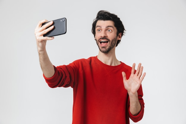 Image of a handsome young excited man posing over white wall take a selfie by mobile phone talking waving.