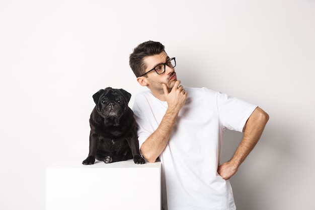 Image of handsome young man, pet owner standing with his dog and looking right at logo, posing with pug against white background