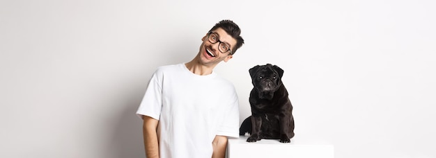 Image of handsome young man standing near cute black pug and smiling dog owner spending time with hi