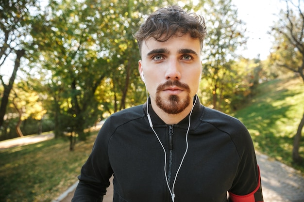 イヤホンで音楽を聴いている公園で屋外でハンサムな若いスポーツフィットネス男ランナーの画像。