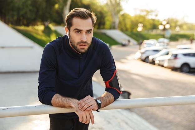 Image of a handsome young strong sports man posing outdoors at the nature park location listening music with earphones.