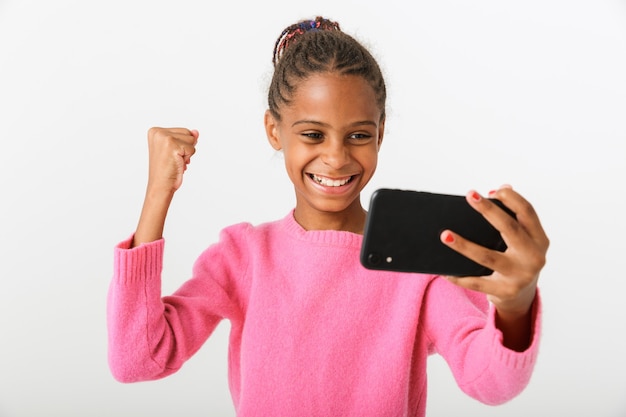 Image of happy african american girl playing video game on cellphone and showing winner gesture isolated over white wall