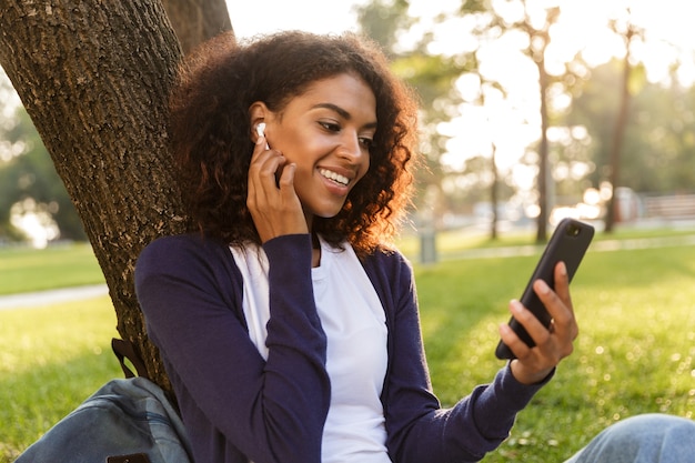 Image of happy african young woman sitting outdoors in park using mobile phone listening music with earphones.