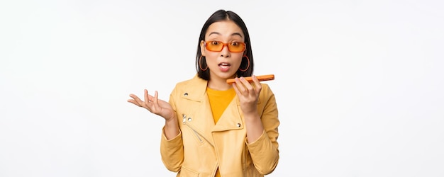 Image of happy asian girl talking on speakerphone recording translating her voice with mobile phone app talking in smartphone dynamic standing over white background