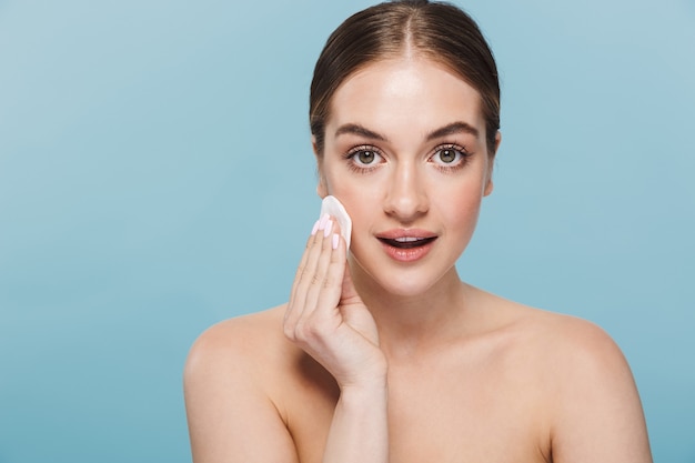 Image of a happy beautiful young woman take care of her skin isolated over blue wall with cotton pad.