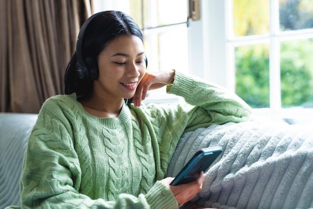 Image of happy biracial woman in headphones using smartphone sitting on sofa at home, copy space. Leisure, relaxation, technology and domestic life concept.