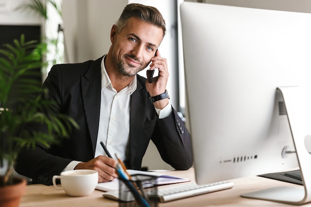 Image of happy businessman 30s wearing suit talking on cell phone while working on computer in office