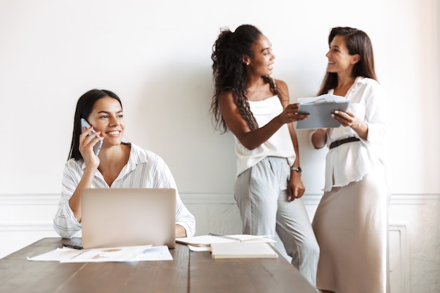 Image of a happy cheery young business woman at workplace indoors using laptop computer talking by mobile phone with blurred colleagues